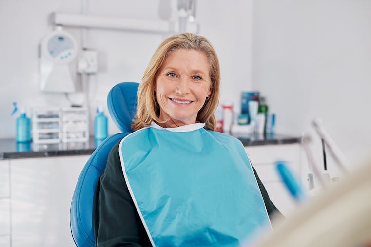 Old beautiful lady smiling while seated on dental chair wearing a blue bib for dental implant. Confident elderly patient waiting for whitening treatment in clinic, dentures and dental prosthesis.